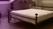 A neatly made metal-framed bed with white bedding is situated in a room with wooden floors and a slanted ceiling. There is a chair to the left and a lamp in the corner.