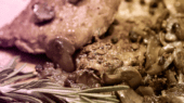 Close-up of a cooked dish featuring slices of meat with mushrooms and a sprig of rosemary on the side, all placed on a wooden surface.