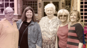 Five women standing together and smiling in front of a brick building with windows and a black fence.