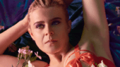 A person with short blonde hair lies on a dark floral sheet, wearing a red outfit and surrounded by white flowers. They look off into the distance with a thoughtful expression.