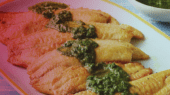 A plate of five cooked fish fillets topped with green sauce, placed on a white and yellow plate. There is a bowl of additional green sauce in the background on a blue tablecloth.