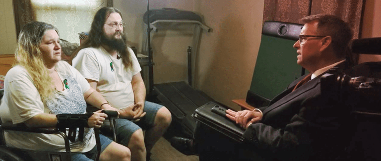 A man in a suit interviews a seated couple in casual attire. The couple is sitting close to each other in a room with a treadmill and a green chair in the background.