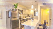 Modern kitchen with stainless steel appliances, white countertops, and an island with two bar stools. Cabinets are light-colored, and two pendant lights hang above the island.