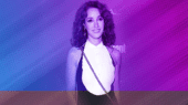 A woman with curly hair, wearing a sleeveless white and black dress, smiles while posing in front of a purple and blue gradient background. She carries a bag with a chain strap over her shoulder.