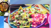 A platter of nachos topped with melted cheese, ground meat, avocado, and diced tomatoes, with a pan of cooked meat and vegetables in the upper left corner.