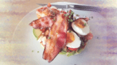 A dish containing avocado, halved boiled eggs, tomato pieces, and two crispy bacon strips, all garnished with chopped chives, served on a white plate with a knife.