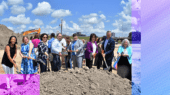 A group of people in business attire participate in a groundbreaking ceremony, each holding a shovel and posing in front of a dirt mound with construction equipment visible in the background under a blue sky.