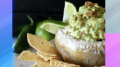 A bowl of guacamole garnished with lime wedges and surrounded by tortilla chips. Two whole jalapeños are in the background.