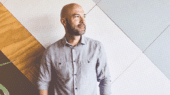 A man with a bald head and beard wearing a gray button-up shirt stands in front of a geometric-patterned wall, looking to his left.