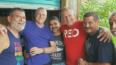 A group of six men, casually dressed, smiling and standing closely together in front of a bar setting with plants in the background.