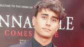 A person with short, styled hair wearing a dark outfit stands in front of a backdrop with text partially visible, including "Annabelle Comes Home.