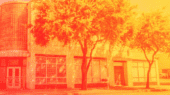 A two-story building with large glass windows on the ground floor and trees lining the sidewalk in front. The photo is tinted with an orange hue.