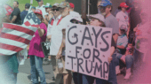 A group of people at a rally holding various signs, including one that reads "Gays for Trump" and another with an American flag design.