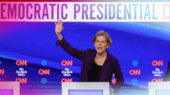 Three individuals stand behind podiums during a Democratic presidential debate hosted by CNN and The New York Times. The person in the center is raising their hand while speaking.