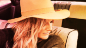 A person with wavy blonde hair is wearing a wide-brimmed hat and a dark shirt, seated in a car with beige upholstery.