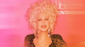 Person with curly, platinum blonde hair and a smile, wearing a black outfit with a pink backdrop.