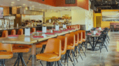 A modern restaurant interior with orange bar stools along a counter, tables with black chairs, menus, and table settings. The kitchen is visible in the background.