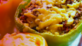 Close-up of a stuffed bell pepper topped with melted cheese, filled with a mixture of ground meat and vegetables, placed on a metallic surface with a partially visible yellow bell pepper nearby.