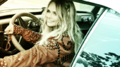 A woman with long blonde hair, wearing a studded jacket, sits in the driver's seat of a vintage car, smiling and looking to her left.