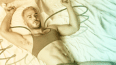 A person with short blond hair lies on a bed, wearing a grey sports bra. Their arms are raised above their head, and there are artistic lines resembling wings drawn around them.