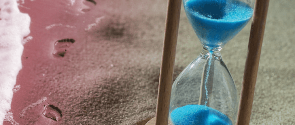 An hourglass with blue sand is placed on a sandy beach next to footprints near the water’s edge.