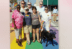 A group of people and a dog stand on a rainbow crosswalk. They are dressed in casual clothing and some wear rainbow-themed items. Another person is seen walking in the background.