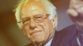 An elderly man with white hair and glasses is wearing a suit and pointing forward.