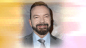 A man with dark hair and a beard in a grey suit and striped tie smiles at the camera against a background with a yellow and pink gradient.