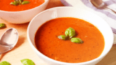 Two bowls of tomato soup garnished with basil leaves, placed on a wooden surface with a spoon and a striped napkin beside them.