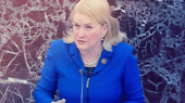 A person with blonde hair in a blue jacket speaks at a podium in front of a marbled wall.